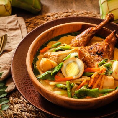 a bowl of curry noodles with chicken and vegetables. The table is decorated with curry leaves, cutleries and two ketupat. Buffet.