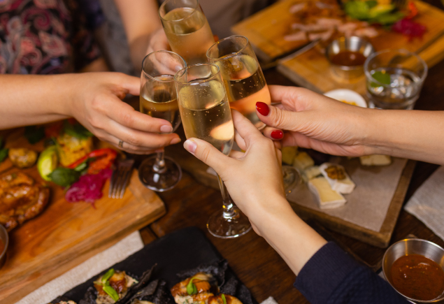 The photo shows four hands holding a champagne clinking their glasses together. The background is a table full of food.
