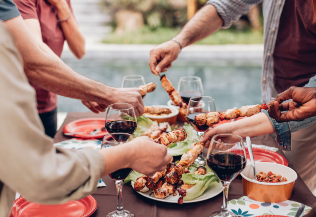A group of friends are holding barbecue sticks with glasses of wine.