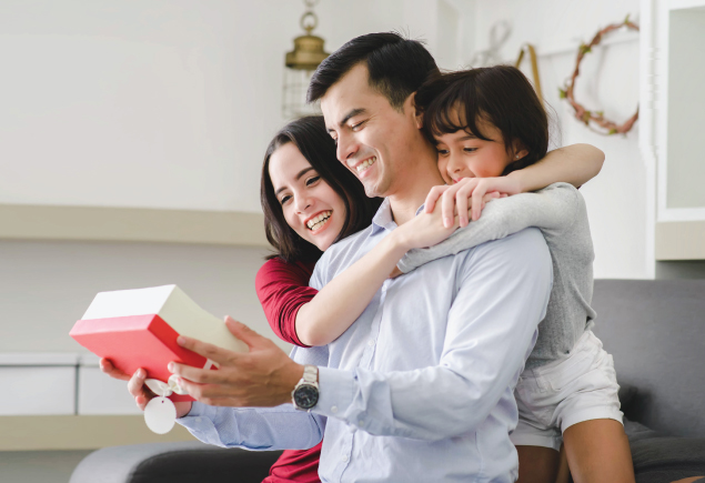A family of three is celebrating Father's Day. The mother and daugther is hugging the father. The father is holding the present looking at it.