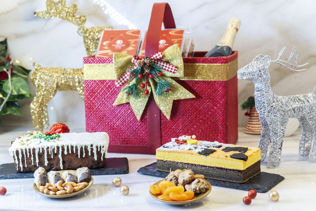 The gift basket consist of a bottle of wine, fruit cake and assorted cookies. The surrounding has Christmas decorations.