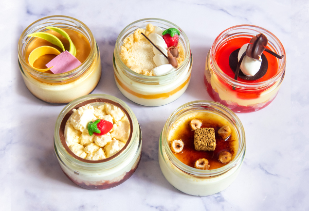 Five desser jars such as creme brulee, coconut pineapple desire, fruit trifle and creme caramel on the table.