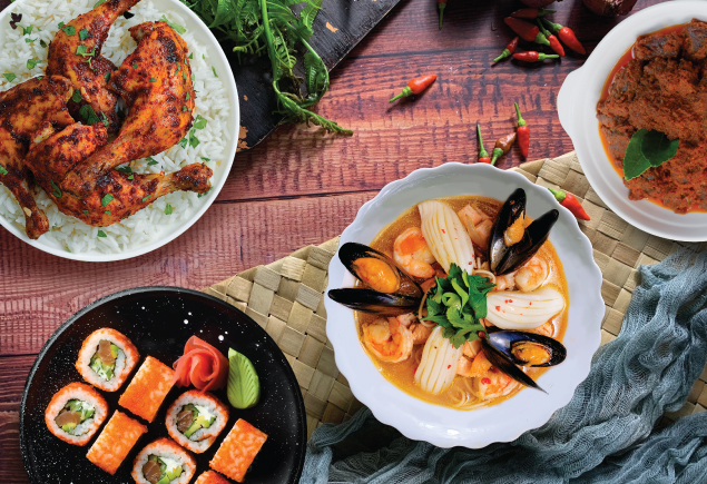 A flatlay photo of a variety of Asian food on the table. From the right, it is a plate of rendang, a bowl of tom yum soup, sushi and ayam penyet with white rice on top left of the photo. The table is decorated with chilis, cloth, paku pakis and rattan mat.
