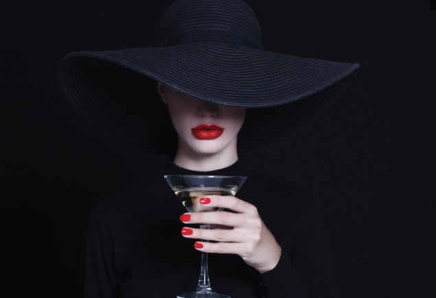 A photo of a lady wearing a black hat covering half of her face and holding a glass of cocktail. She is also wearing a red lipstick and nail polish.