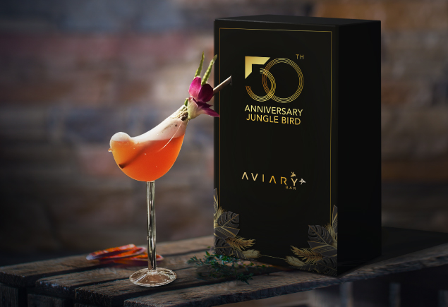 A glass of the jungle bird cocktail in a bird-shaped glass. The 50th anniversary box for the glass is placed right behind the cocktail.