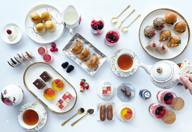 A flatlay photo of a high tea set. Featuring sweet and savoury finger bites such as scones, eclair, cakes, cake pop, jams, berries, sandwiches, burgers, tarts and pastries.