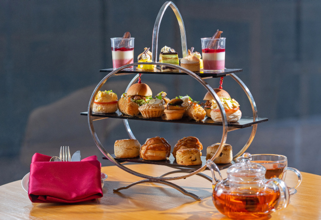 A three-tier high tea set with a pot and cup of tea at the side. The high tea set consists of various sweet and savoury finger bites such as cakes, scones, burgers, and sandwiches. The high tea, tea, and cutleries wrapped in a napkin are all placed on a wooden table.