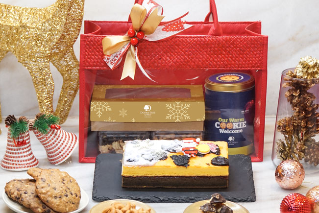 The photo shows a few Christmas desserts such as fruit cake and biscuits. There is a red packaging bag behind the fruit cake. In the bag, it consist of a can of DoubleTree cookies, fruit cake and assorted biscuits.