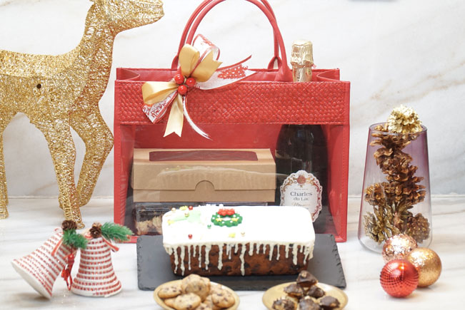 The photo shows a red bag that consist of fruit cake, assorted biscuits and a bottle of wine. At the front of the bag, there is a fruit cake and biscuits. The surrounding is decorated with Christmas decorations.