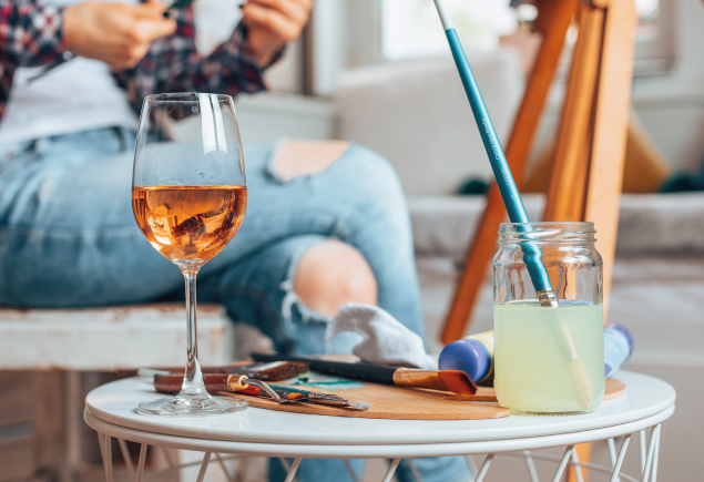 A glass of wine, a few paint brushes and watercolor are on a table.