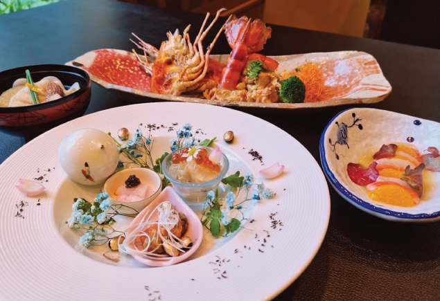 The photo shows four Japanese cuisine. The left dish is a Chicken Broth with Sea Cucumber and Clam Soup. The dish at the bottom Abalone with Yuzu Jelly and the dish on top is Teppan Grilled River Prawn with Miso Flavored Butter Sauce. The right dish is Hokkaido Scallop and Salmon Carpaccio.