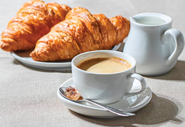 The photo shows a cup of hot coffee with one biscuit on the side, a small jug of fresh milk and two pieces of croissant.