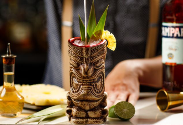 Aviary Bar's signature cocktail, Jungle Bird. The cup is made of wood and it has a face crafted on it. The background shows a mixologist preparing for another cocktail with the ingredeints on the table.