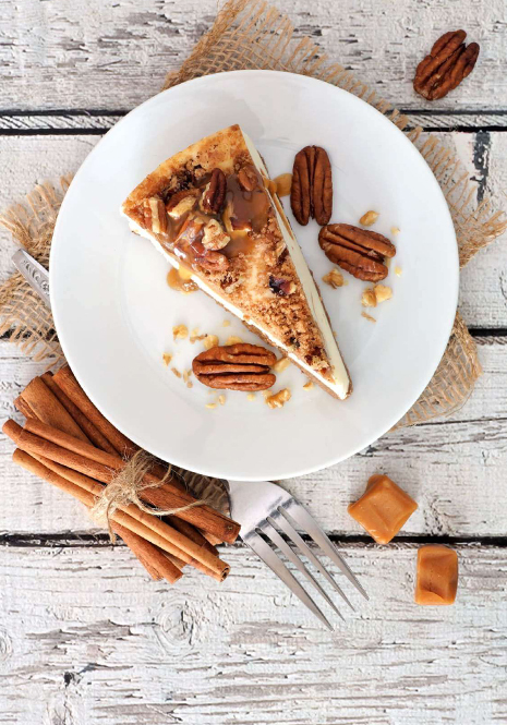 A slice of pecan cake is on a plate. The wooden table is decorated with pecan nuts, cinnamon sticks and a fork.