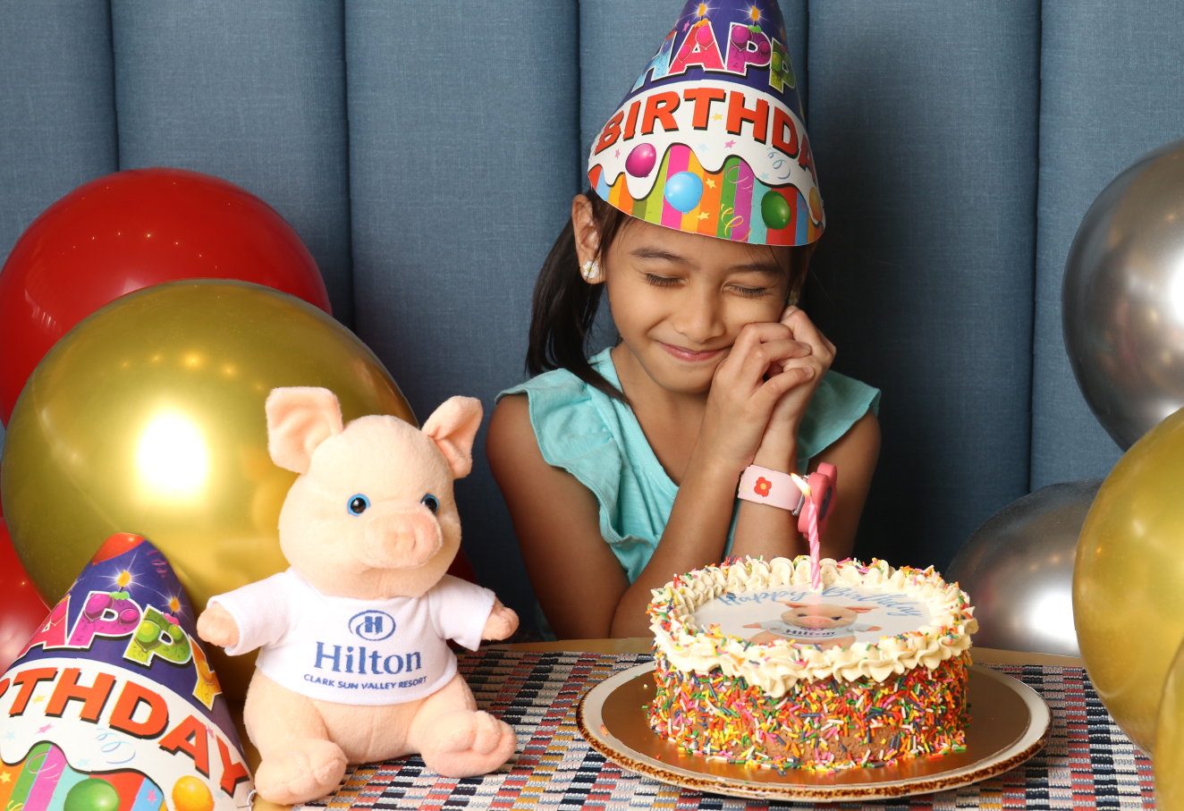 A girl celebrating her birthday. She is wearing a birthday hat and making a wish. The table has a birthday cake with one candle, a piglet plushie, and a few balloons.