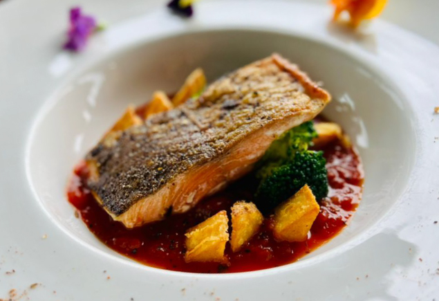 A plate of pan-seared salmon filet with livornese sauce. It is also served with roasted potatoes and broccoli.