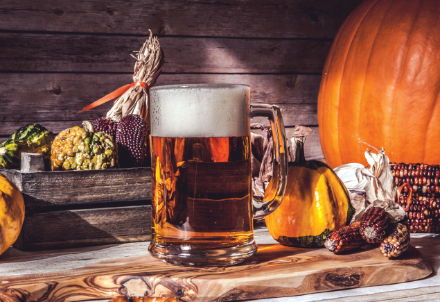 The photo shows a mug a beer placed on top of a wooden table. The table also has various items such as corns, broccoli, pumpkin and an ornamental glass pumpkin.