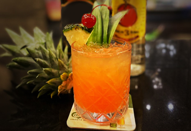 A glass of the Jungle Bird cocktail with pineapple, cherry and pandan leaf decorated on top.