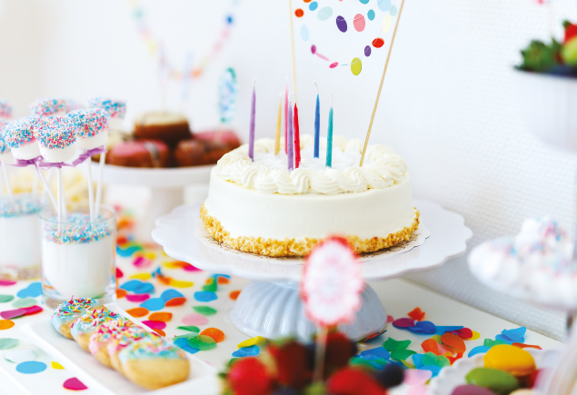 A birthday cake with seven sticks of colourful candles on top of it. The background is decorated with a variety of sweets treats and colourful confetti.