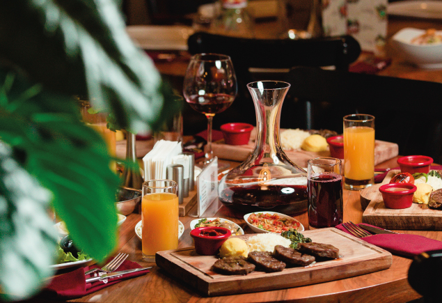 A variety of food, a glass of wine and three glasses of juices are on the table.