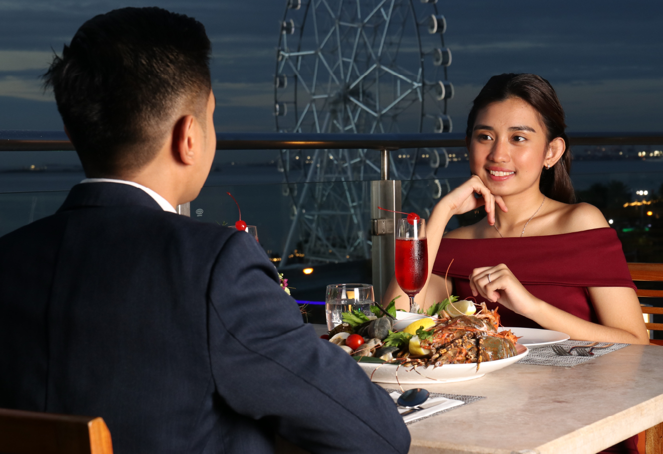 The photo shows a couple looking at each other. The man's back is facing the camera and the woman is looking at the man. Both of them are dressed in formal attire; the man is wearing a dark blue blazer and the woman is wearing an off-shoulder red dress. On the table, there is a plate of fresh seafood and a few glasses of drinks.