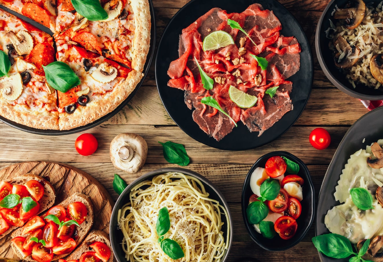 Six different Italian dishes are on a wooden table. There is pepperoni pizza, cured meat, mushroom risotto, ravioli, spaghetti and a few slices of sourdough bread with tomatoes on top.