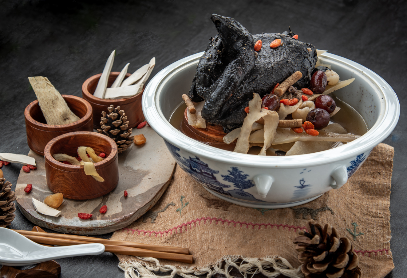 Eight Treasure Herbal Soup with whole Black Chicken in a huge bowl. Three bowls of herbal ingredients on the side.