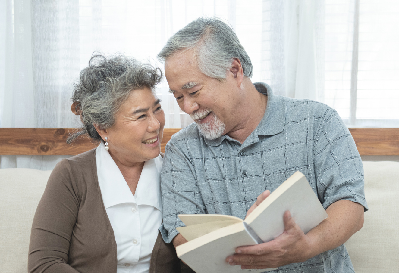 A pair of elderly asian couple is sitting very close together showing affection to each other. The elderly asian man is holding and pointing to the book's contents while looking and smiling at the woman. The woman is seated close to the man and looking at him smiling.