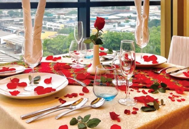 Two plates, two set of utensils, a few glasses on the table decorated with rose petals that sets up a romantic ambience.