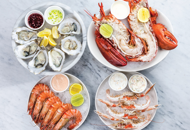 Four different types of fresh seafood are placed on a marble table. From the top left it's a plate of fresh oysters and beside it is a plate of lobsters. On the bottom left it's a plate of prawns and beside it is a plate of crab legs.