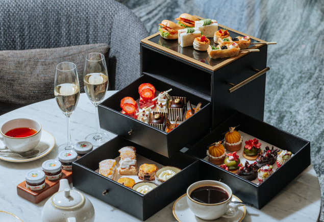 An afternoon tea set for two person. It has a variety of desserts, sandwiches, a cup of tea and coffee and two glasses of champagne.