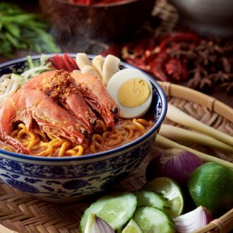 A bowl of Penang Prawn Noodles decorated with three prawns, half hard boiled egg, and other condiments on top.