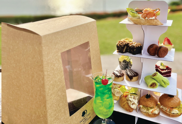 The photo shows a high tea set for two with a glass of drink on the table. There is a takeaway box placed beside the high tea set. The photo was taken outdoor with the seaside as the background.