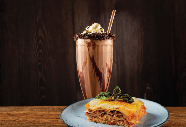 A glass of chocolate milkshake and a plate of lasagna.
