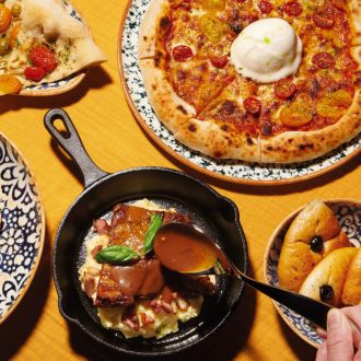 A flatlay photo of Italian cuisine such as pizza, bread and brownie dessert are on a wooden table. A person is holding a spoon to drizzle the brownie.
