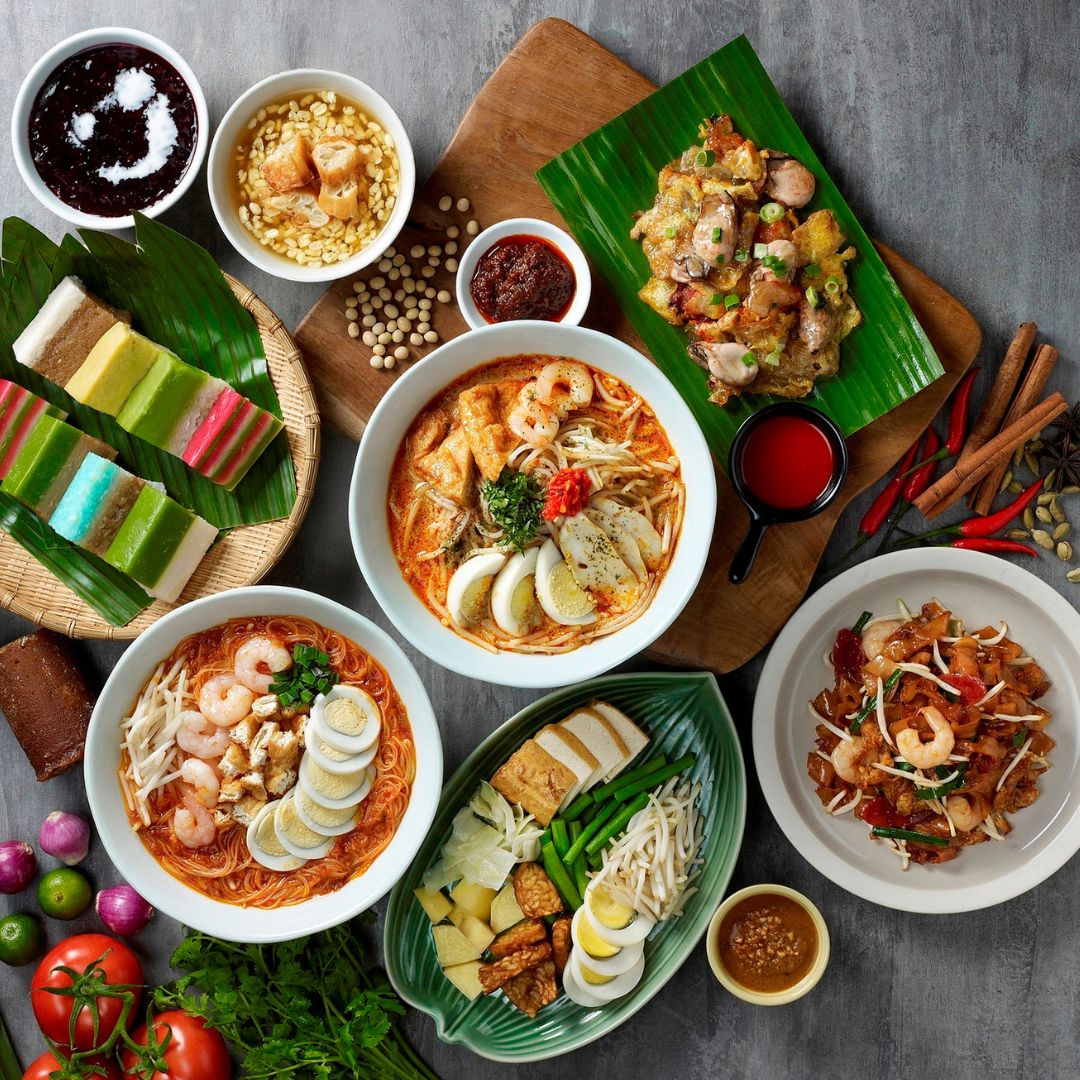 The photo shows six types of Asian cuisine such as Curry Noodle, Prawn Noodle, Assorted Malay Kuih, Char Kuey Teow and Oyster Omelette. There are five different types of sauces and three types of condiments on the table.