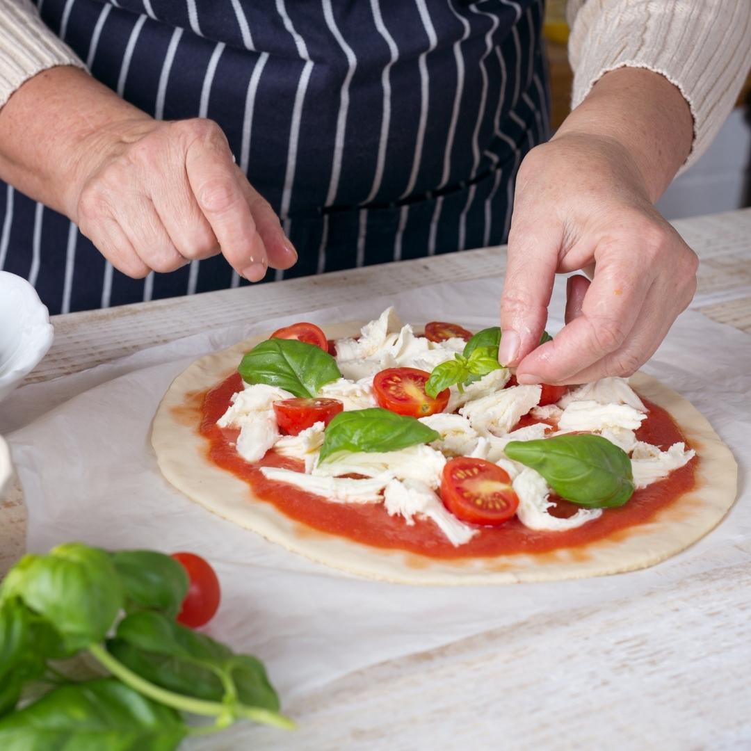 A chef is preparing a Margherita Pizza on thin white cloth that is placed on a wooden table. He is preparing all the ingredients on the pizza dough. The ingredients used are tomato sauce, sliced cherry tomatoes, mozzarella and basil leaves. A bunch of basil leaves and a piece of cherry tomato are blurred out on the side of the photo. The chef is wearing a dark blue with white striped apron and a cream coloured sweater.