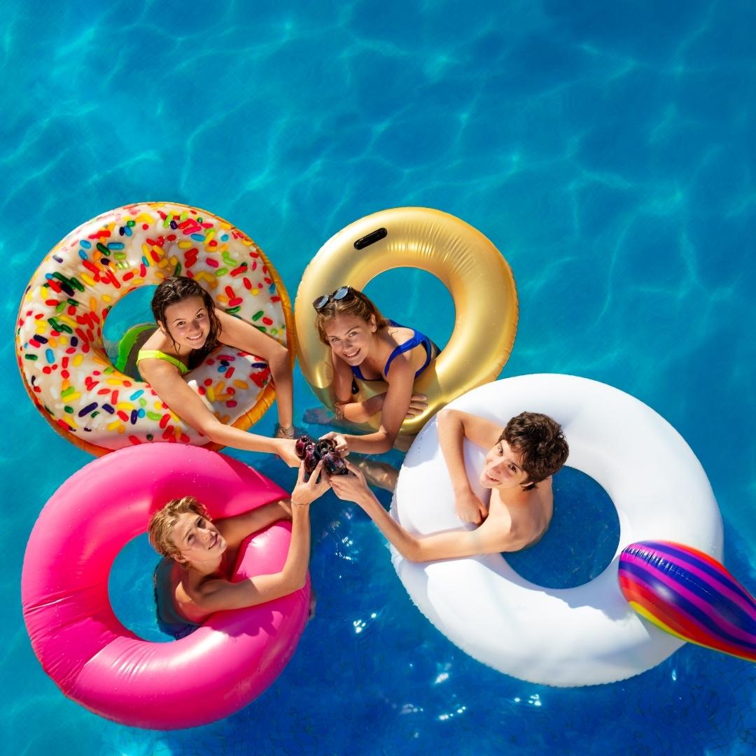 A group of friends are looking up and clinking their drinks to pose for a photo in the pool. There are two females and two males, each of them has a pool float. From top left, the girl has a sprinkled donut float, the girl on the right has a gold float, the boy on the bottom left has a hot pink float and the boy on the right has a unicorn float.