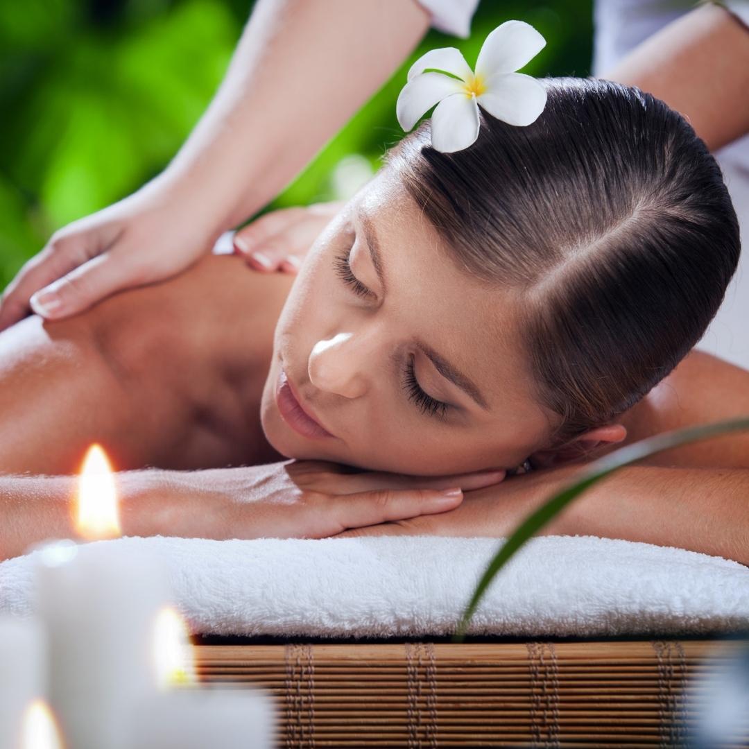 A women is lying down on her front with her hands supporting her head. She is getting a back massage in a spa. Her hair was tied neatly with a stalk of white flower tucked on the top part of her head. The spa has a few candles lit up to have a relaxing atmosphere.