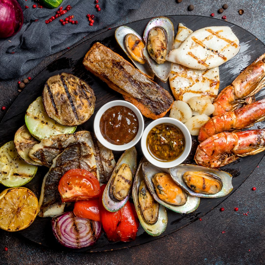 The photo shows a variety of grilled seafood and vegetables such as prawns, mussels, squid, salmon, aubergine, tomatoes, onions, and cucumbers.