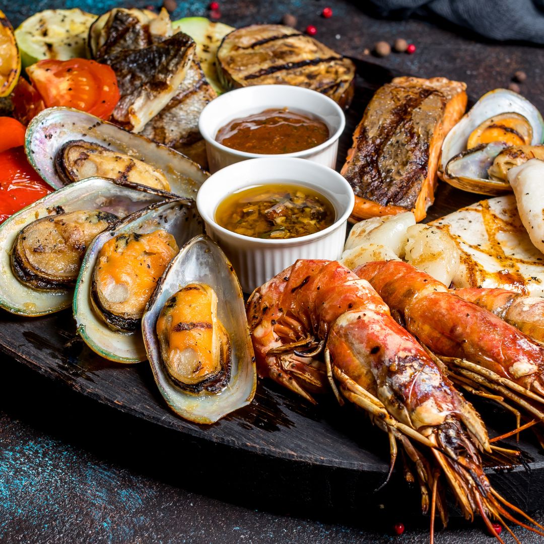 The photo shows a plate of assorted grilled seafood such as prawns, mussels, salmon, and squid. It also includes grilled vegetables such as tomatoes, cucumbers and aubergine.