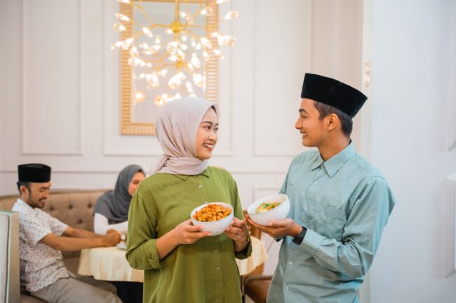 A group of four friends are breaking fast together. A Muslim woman and a Muslim man are standing in front of the camera looking at each other; each holding a bowl of food. The other two friends are at the back sitting down at the dining table.