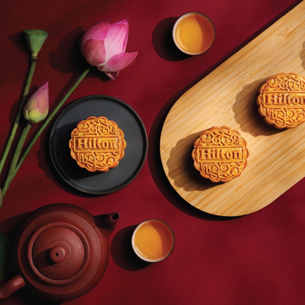 Three pieces of Hilton baked mooncake. Decorated with a pot of tea, two cups of tea and lotus flower.