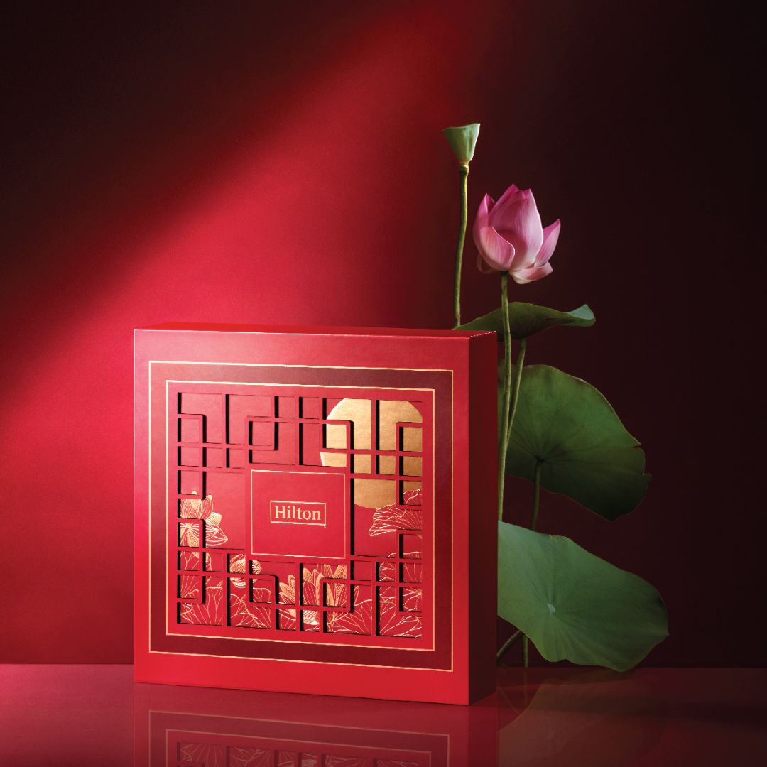 Hilton Malaysia's 2023 mooncake national box. The box is red in colour and oriental designs are imprinted on it. Beside the box, there is a lotus flower.