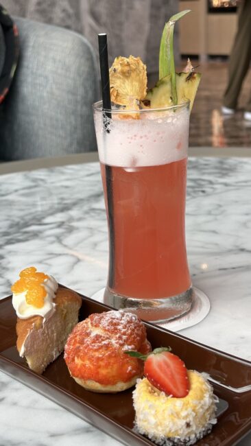 A glass of mocktail and a tray of 3 cakes beside it.